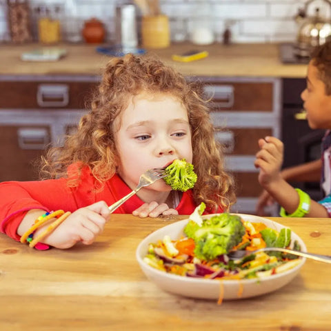 A young child with curly hair enjoys broccoli from a fork at the kitchen table, their veggies topped with Gustus Vitae Kid’s Menu: Veggie: All Purpose Seasoning, while another child is in the background. A bowl of salad sits in front of them.