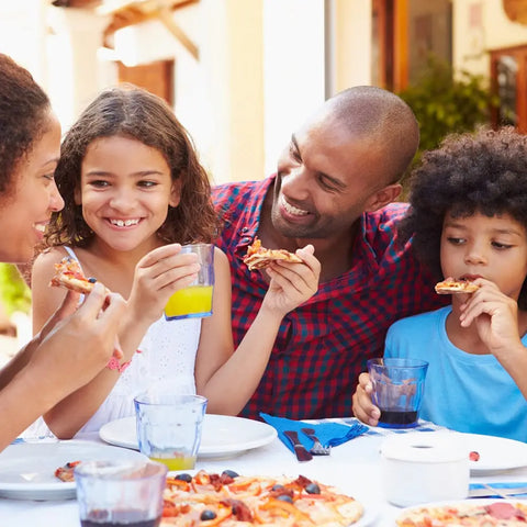 A family of four enjoys pizza topped with Gustus Vitae Kid’s Menu: Veggie: All Purpose Seasoning. The mother and kids hold slices as the father smiles at his daughter. Drinks and non-GMO foods are on the table.