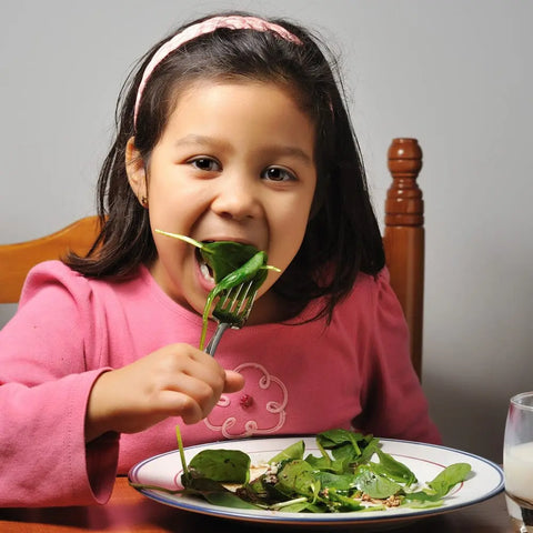 A young girl in a pink shirt smiles at the table, enjoying fresh spinach from her salad topped with Gustus Vitae Kid’s Menu: Veggie: All Purpose Seasoning, made from non-GMO ingredients, next to a glass of milk.