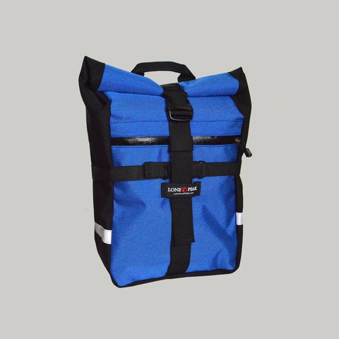 The Lone Peak Packs Kings Peak Waterproof Pannier is a blue and black roll-top backpack featuring a front zip pocket, black strap and buckle, reflective strip, adjustable pannier hooks, top handle, and the Lone Peak logo on the front.