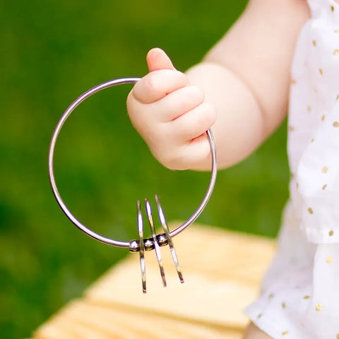 A baby in a white shirt with gold dots holds the Kleynimals Stainless Steel Rattle by Kleynimals, featuring three smaller rings, outdoors against a blurred green background.