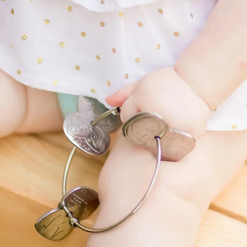 A baby in a white outfit with gold polka dots holds a Kleynimals Stainless Steel Rattle by Kleynimals, featuring heart-shaped charms, while sitting on a wooden surface.