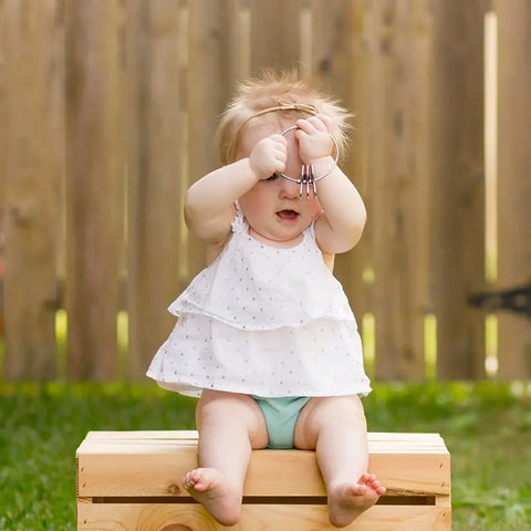 A baby in a white dress sits on a wooden box outside, holding the Kleynimals Stainless Steel Rattle by Kleynimals above their head, with a wooden fence and grass in the background.