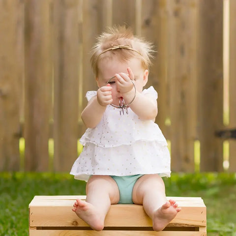 A baby in a white dress and teal diaper cover sits on a wooden box outdoors, holding the Kleynimals Stainless Steel Rattle by Kleynimals. A wooden fence and grass are visible. This non-toxic, eco-friendly rattle makes a perfect baby gift.