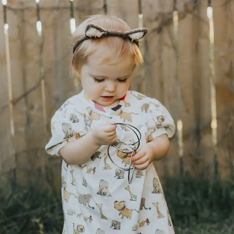 A toddler in an animal print dress and cat ear headband stands by a wooden fence, looking down and playing with Kleynimals Stainless Steel Jangles® by Kleynimals among the metal bangles in their hands.
