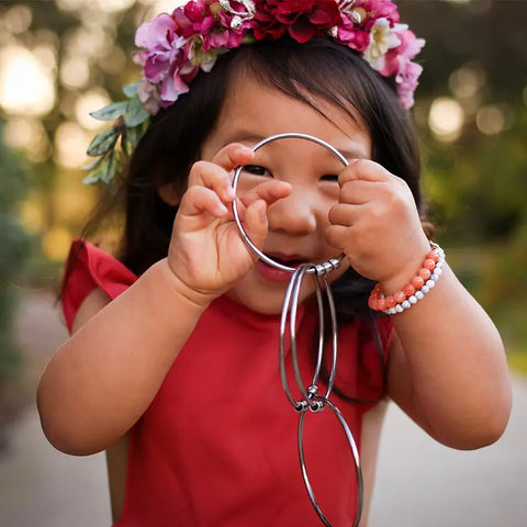 A young girl in a red dress and floral crown smiles while holding the Kleynimals Stainless Steel Jangles® teether by Kleynimals. She wears orange and white beaded bracelets and stands outdoors in soft, natural light.