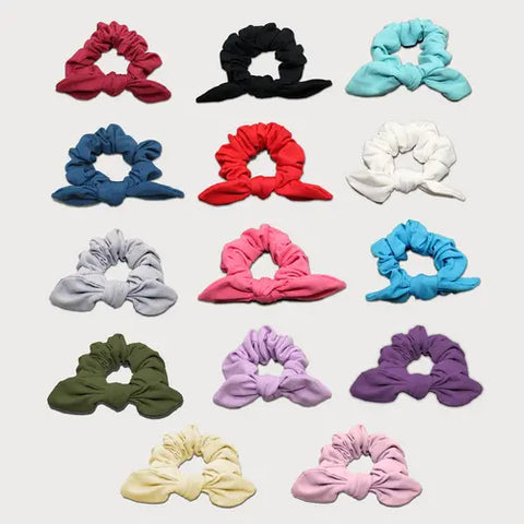 A set of 15 Goodnighties Knotted Bow Scrunchie hair accessories in assorted colors—red, black, blue, purple, pink, green, white, and yellow—displayed in a 5x3 grid on a white background. Made in the USA.