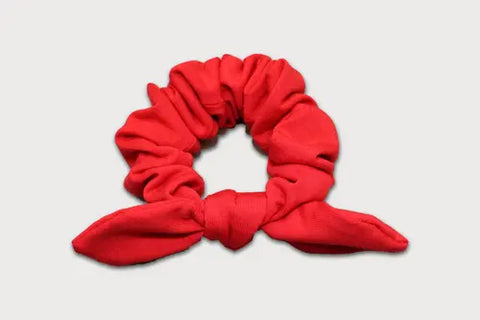 The Goodnighties Knotted Bow Scrunchie, in bright red thermoregulating fabric with two pointed bow ends, is displayed on a plain light grey background.