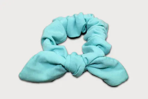 The Goodnighties Knotted Bow Scrunchie, crafted from thermoregulating fabric in light blue with two fabric ends tied in a bow, is displayed against a plain white background.
