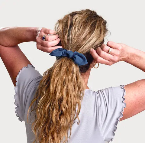 A woman with wavy blonde hair ties her hair into a ponytail using the blue scrunchie from the Goodnighties Matching Knotted Bow Headband & Scrunchie set. She wears a light gray, short-sleeved top with ruffled edges and rings against a plain background.