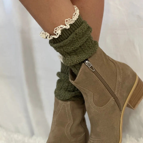 A close-up shows a persons legs wearing Catherine Coles BOOTIE Lace Slouch Socks in olive, featuring white lace trim, styled with light brown suede ankle boots with side zippers and low wooden heels against a plain white background.