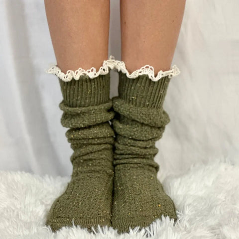 A pair of feet wearing Catherine Cole BOOTIE Lace Slouch Socks in olive with white trim stands on a soft, white fuzzy surface against a white background.