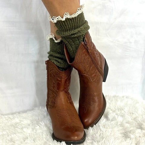 A person stands on a fluffy white carpet wearing brown leather Western ankle boots and Catherine Cole BOOTIE Lace Slouch Socks in olive, featuring white lace trim.