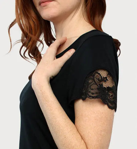 A woman with wavy red hair models the Goodnighties Lace Accent Scoop Neck Tee in black, shown from the side with one hand on her collarbone against a plain light background.