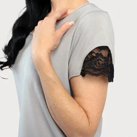 A person wears the Goodnighties Lace Accent Scoop Neck Tee, a light gray top with black lace trim on the sleeve, and holds their hand near their neck against a plain white background.