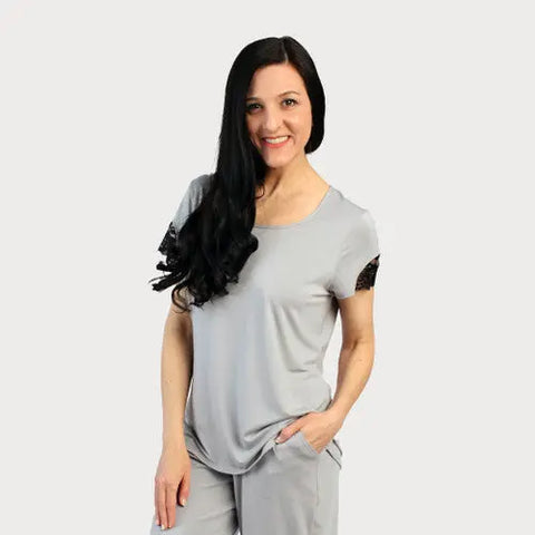 A woman with long dark hair smiles, wearing the Goodnighties Lace Accent Scoop Neck Tee in light gray and matching pants, posing with one hand in her pocket against a plain white background.
