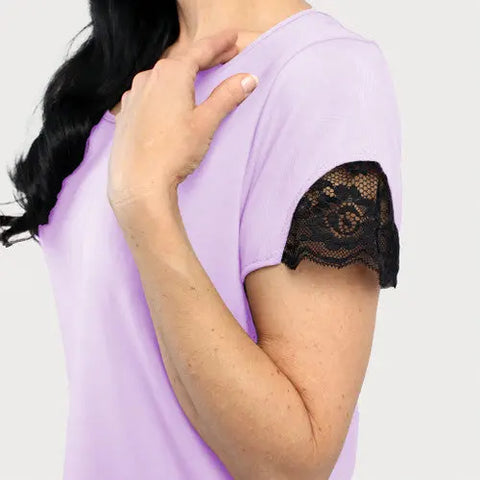A woman wears the Goodnighties Lace Accent Scoop Neck Tee in light purple, featuring black lace trim on the sleeves. She touches her shoulder with one hand against a plain, light-colored background.