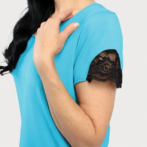 A woman wears the Goodnighties Lace Accent Scoop Neck Tee in turquoise with black sleeve trim, touching her collarbone. Only her upper body is shown against a plain light background.