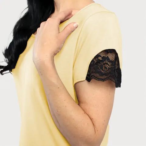 A person with long dark hair wears the Goodnighties Lace Accent Scoop Neck Tee, a short-sleeved yellow top with black lace sleeve trim, holding their hand near their shoulder against a plain background.