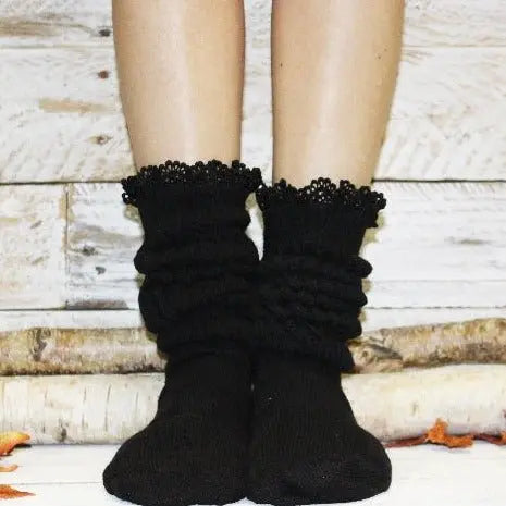 A pair of legs in Catherine Cole’s LACE Topped Ultimate Slouch Socks in black, standing on a white surface with wood pieces and orange leaves—ideal chic womens fashion accessories.