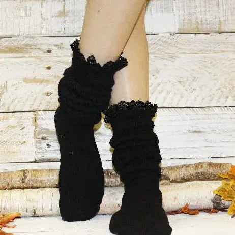 A person models Catherine Cole’s LACE Topped Ultimate Slouch Socks in black, standing with crossed legs on a rustic wooden floor strewn with autumn leaves against a whitewashed wood backdrop.