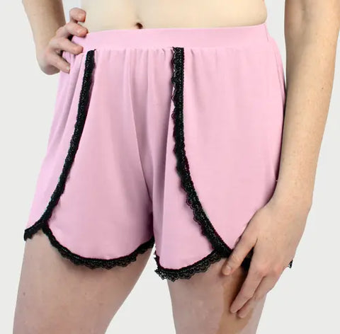 A person models Goodnighties Lace Trim Tulip Shorts—stylish American-made sleepwear in light pink with black lace trim—standing with one hand on their hip against a plain light background.