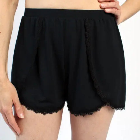 A person wearing Goodnighties Lace Trim Tulip Shorts—American-made sleepwear in black—stands with one hand on their hip against a plain background.