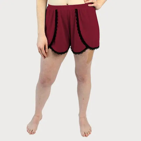 A person stands barefoot in burgundy Goodnighties Lace Trim Tulip Shorts, one hand on their hip. Light skin patches are visible against a plain, light-colored background.
