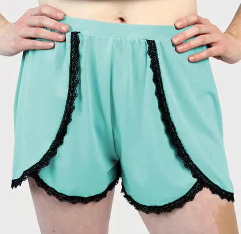 A person stands with hands on hips against a plain background, wearing Goodnighties Lace Trim Tulip Shorts in light turquoise with black lace trim—moisture-wicking loungewear for ultimate comfort and style.