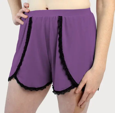 A person stands with one hand on their hip, wearing Goodnighties Lace Trim Tulip Shorts in high-waisted purple, set against a plain background.