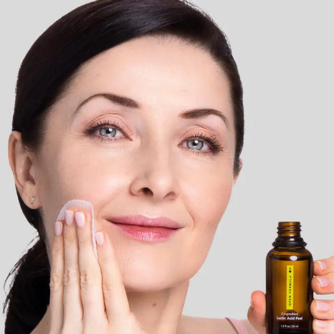 A fair-skinned woman uses a cotton pad to apply Open Formula Lactic Acid Peel to her cheek from a small amber bottle, smiling gently as she enjoys gentle exfoliation. The background is plain light gray.