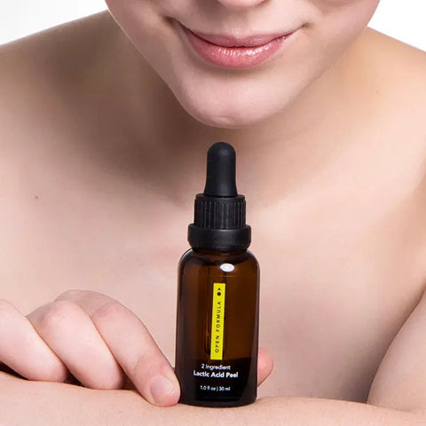 A person with bare skin smiles gently, resting their arm on a surface. In front is an Open Formula Lactic Acid Peel bottle, which exfoliates and unclogs pores. The focus is on the bottle and the persons lower face.