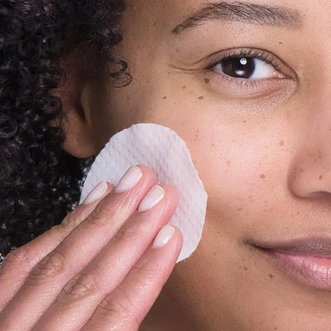 A person with curly hair gently wipes their cheek with a cotton pad, showcasing smooth, glowing skin after using Open Formula Lactic Acid Peel, which exfoliates and unclogs pores for a fresh complexion.