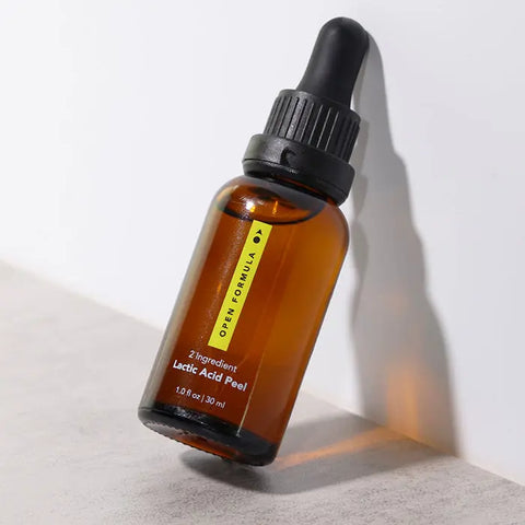 A brown glass dropper bottle labeled Open Formula Lactic Acid Peel rests at a slight angle, casting a soft shadow. This gentle exfoliant from Open Formula uses lactic acid to unclog pores and smooth skin.