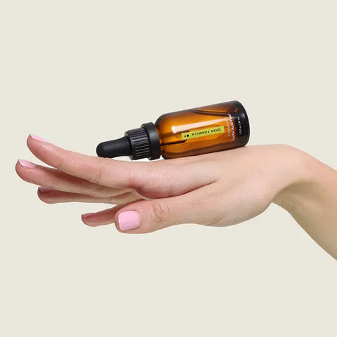 A hand with light pink nail polish is shown palm-down, fingers extended. An amber glass dropper bottle of Open Formula Lactic Acid Peel rests on the back of the hand against a plain light background.