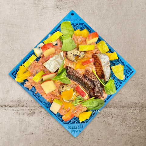 The SodaPup Large Blue Jigsaw & Large Yellow Honeycomb EMat Lick Mat Bundle holds a mix of raw meat, fish, egg yolks, spinach, apple pieces, yellow veggies, and sausage slices on a concrete surface.