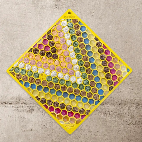 A Large Yellow Honeycomb EMat Lick Mat from SodaPup, part of the Large Blue Jigsaw & Large Yellow Honeycomb Bundle, lies diagonally on a textured surface, with colorful food bits in each cell.