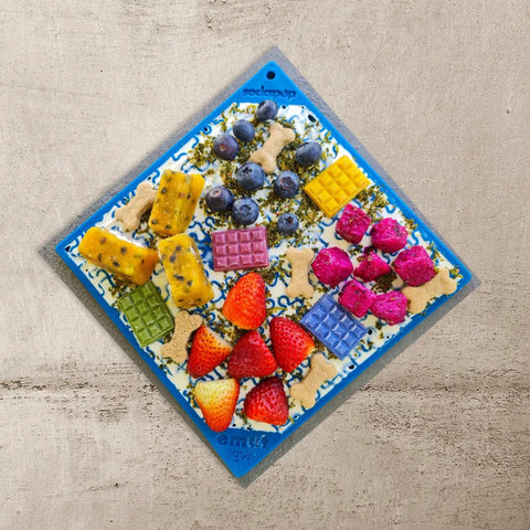 A SodaPup Large Blue Jigsaw EMat Lick Mat from the Large Blue & Pink Jigsaw Bundle is topped with dog treats, blueberries, strawberries, diced yellow fruit, pink dragon fruit, wafer squares, and a creamy spread on a light brown surface.