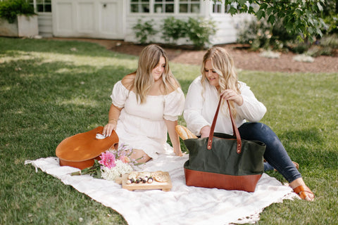 Two women picnic on a grassy lawn. One in a white dress holds a guitar while the other, wearing a white shirt and jeans, looks into her Urban Southern Large Canvas Tote. Flowers and snacks are spread on the blanket.