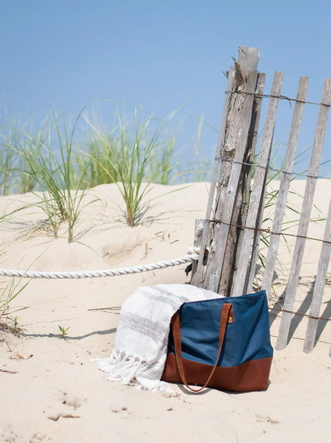 The Urban Southern Large Canvas Tote with full-grain leather accents and a striped towel rest by a weathered wooden fence on a sandy beach, dune grass swaying beneath a clear blue sky.
