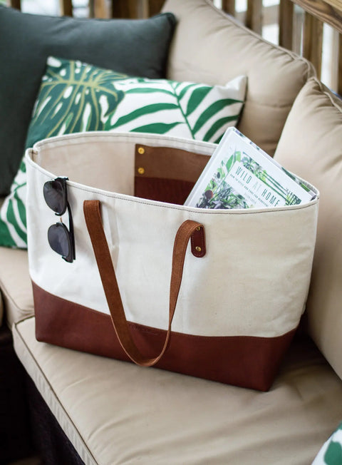 The Urban Southern Large Canvas Tote with full-grain leather accents rests on a beige outdoor sofa. Sunglasses hang from the bag and a copy of Wild at Home peeks out, while leaf-patterned pillows are in the background.