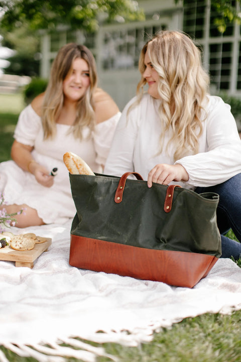 Two women relax on a picnic blanket outdoors. One reaches into an Urban Southern Large Canvas Tote with a baguette inside, while the other smiles holding an item. A wooden board of snacks sits nearby on the blanket.