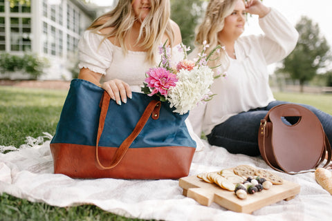 Two women sit on a picnic blanket outdoors. One holds the Urban Southern Large Canvas Tote with full-grain leather accents and flowers, while the other sits nearby. A wooden tray with crackers, cheese, and grapes is in front.