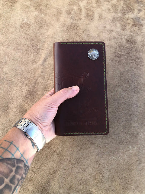 A hand with a tattoo and silver watch holds the Sully + Proper Supply Big Dark Oil Brown Wallet 7 Pocket, American-made and handcrafted in Texas, featuring green stitching and a silver coin detail against a textured tan background.