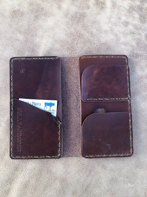 The Sully + Proper Supply Big Dark Oil Brown Wallet 7 Pocket, handcrafted in Texas, features green stitching, multiple card slots and a business card pocket. Shown open on a textured beige surface.