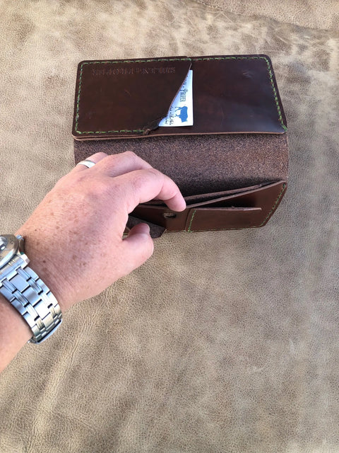 A hand wearing a silver watch opens the Sully + Proper Supply Big Dark Oil Brown Wallet 7 Pocket, handcrafted in Texas, on a tan suede surface, revealing a partially exposed business card inside one of its compartments.