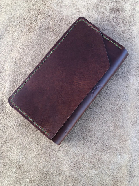 The Sully + Proper Supply Big Dark Oil Brown Wallet 7 Pocket, featuring green stitching and a diagonal front flap, is handcrafted in Texas. It sits closed on a textured tan surface.