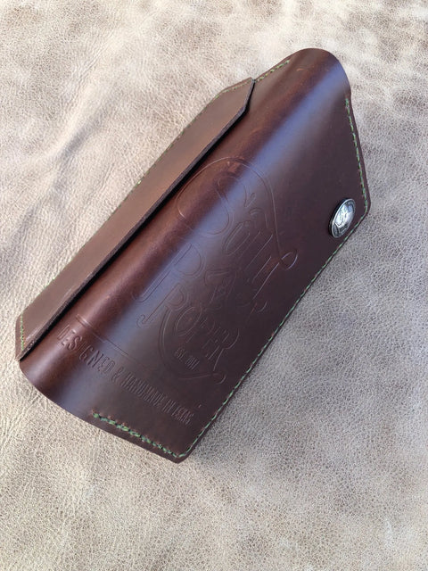 The Sully + Proper Supply Big Dark Oil Brown Wallet 7 Pocket features green stitching, a metal snap button, and South & Roper embossing. Handcrafted in Texas, it sits on a tan textured surface.