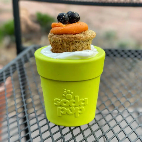 A small dog cake with orange frosting and blueberries sits in a green SodaPup Large Flower Pot Durable PUP-X Rubber ECup Treat Dispenser & Enrichment Toy on a metal table, perfect for dog enrichment.