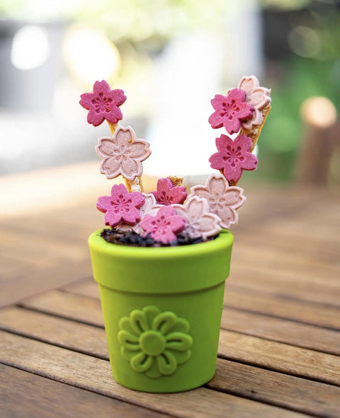 The Large Flower Pot Durable PUP-X Rubber ECup Treat Dispenser & Enrichment Toy by SodaPup holds pink and light pink sakura-shaped cookies on sticks, mimicking cherry blossoms, set on a wooden table with a blurred outdoor backdrop.
